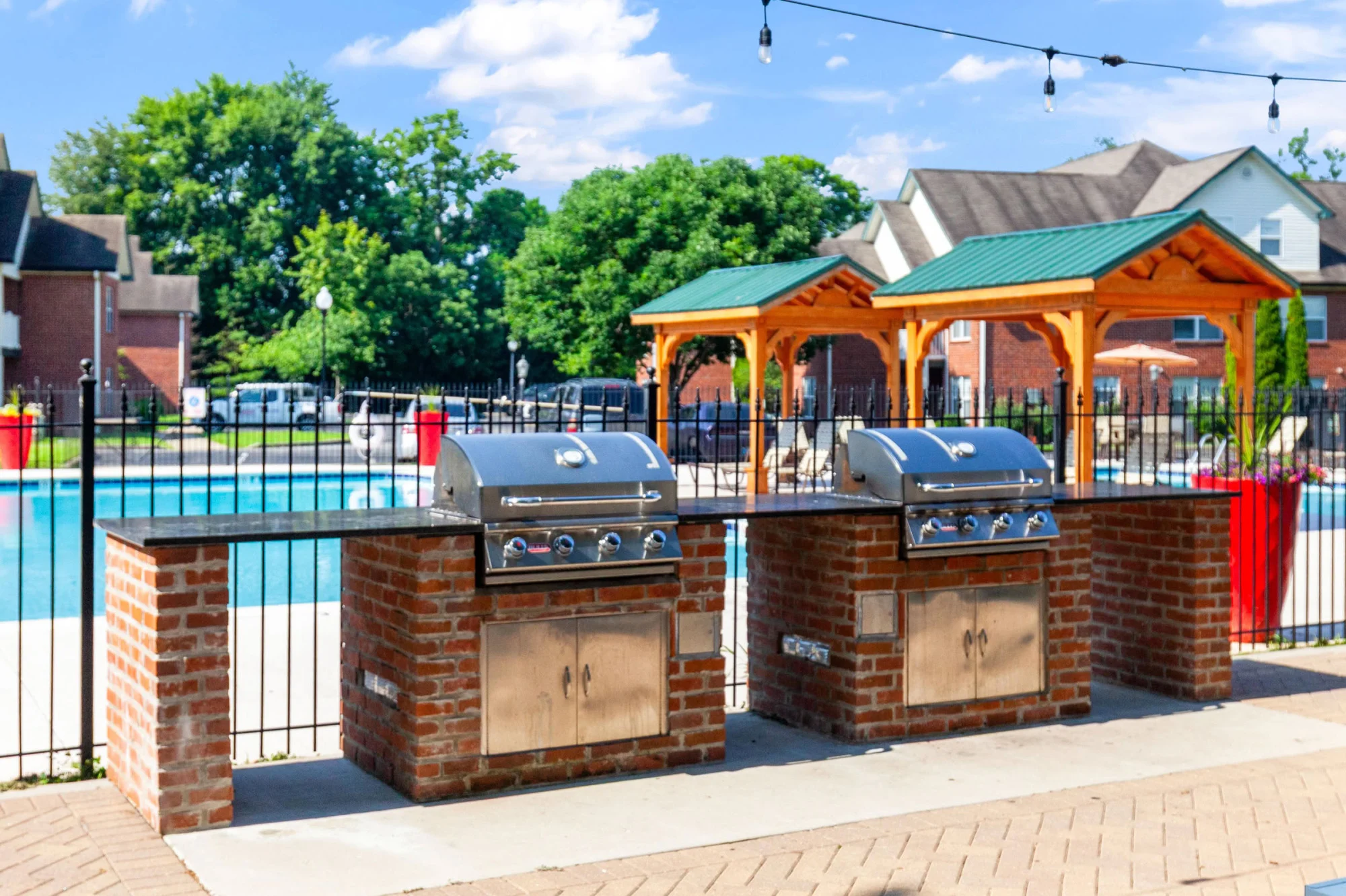 Community outdoor kitchen with two brick-encased stainless steel grills, string lights, wooden pergolas with green roofs, and the swimming pool in the background.