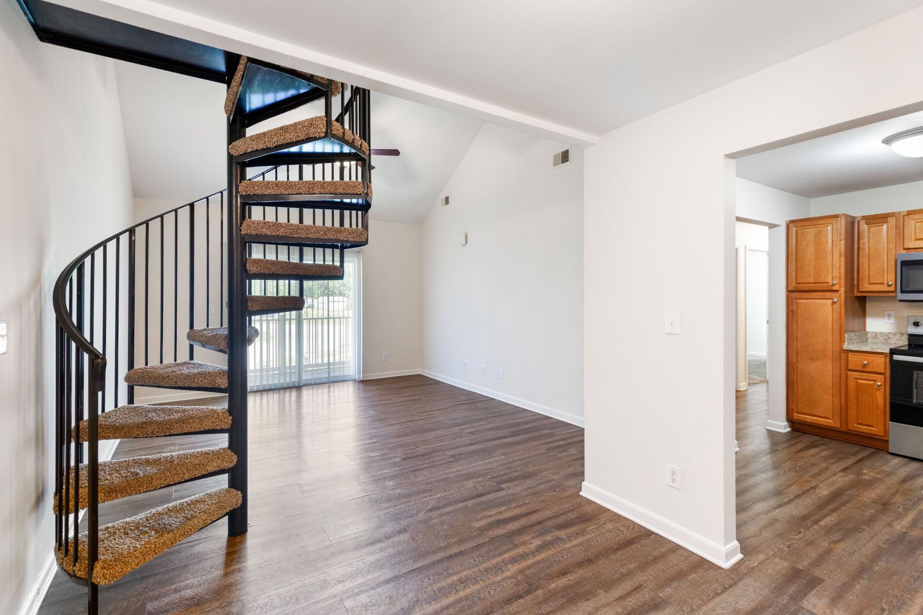 Open-concept apartment loft interior with a black metal spiral staircase, vaulted ceiling, hardwood floors, and a glimpse of the kitchen with wood cabinets and stainless steel appliances.